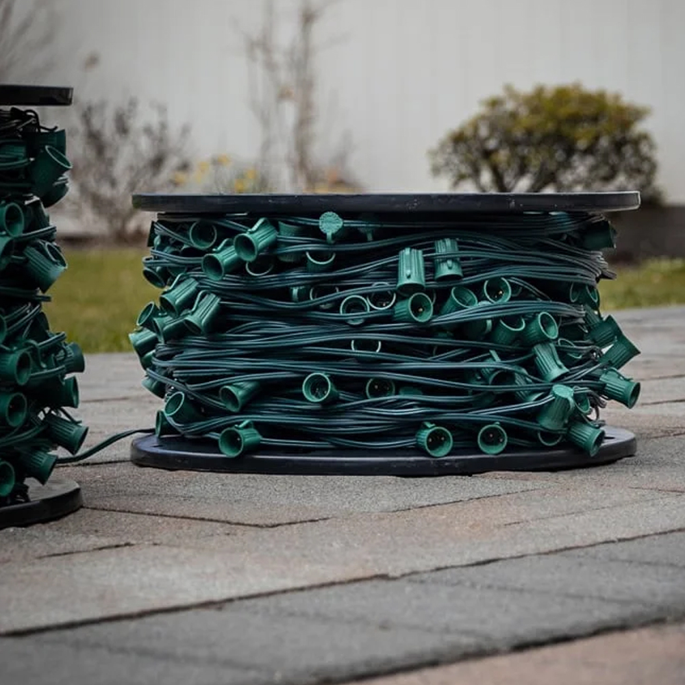 Large spool of bulk C9 light stringer wire with green sockets and wire, set outdoors on a rooftop with a blurred background. C9 Spool 12" Spacing Green 100'