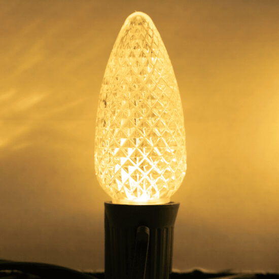 Close-up of a lit C9 LED Christmas bulb in Sun Warm White (deep amber/yellow color) with a faceted, diamond-cut lens. C9 Sun Warm White Faceted Spk